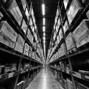 Black and white image of a warehouse aisle filled with stacked boxes and shelves.
